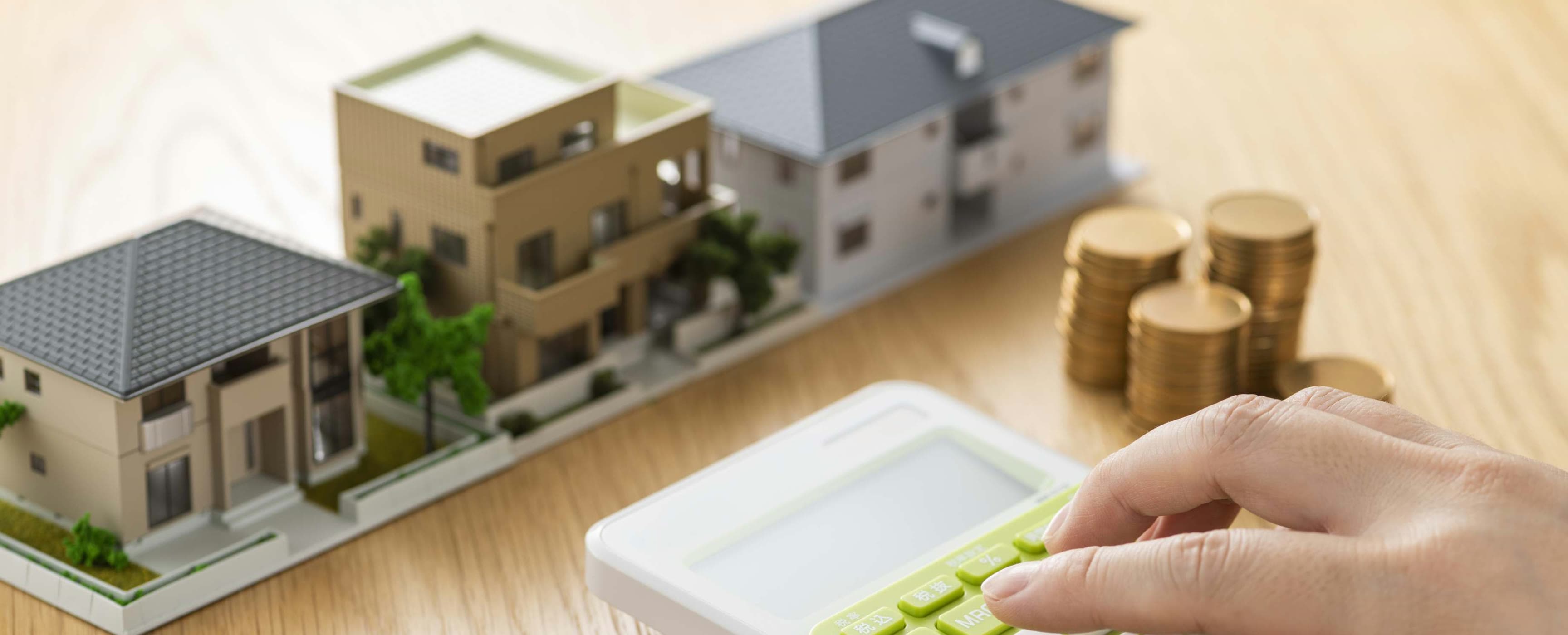 A hand using a calculator beside model houses and stacks of coins